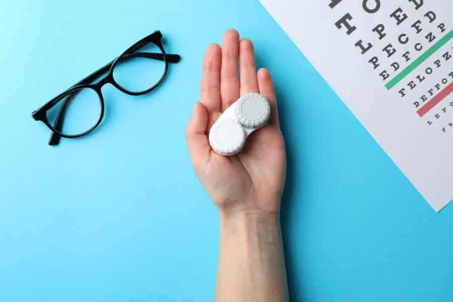 Hand with contact lenses, glasses and eye test chart on blue background, top view stock photo